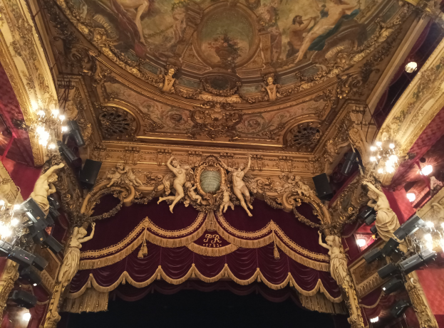 Das Innere eines Theaters mit einem roten Vorhang, Kronleuchtern, Skulpturen, Lichtern, einer Deckenmalerei und einer Bühne mit ein paar Menschen davor.