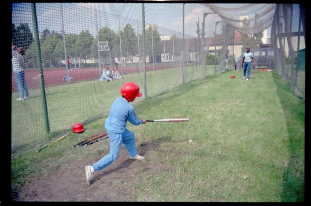 Ein junger Junge in einem blauen Kleid und einem roten Helm schwingt einen Baseballschläger auf einem grünen Feld mit verstreuten Schlägern, Bäumen, Gebäuden und einem klaren Himmel im Hintergrund.