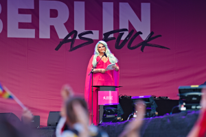 Eine Frau in einem pinken Kleid steht auf einem Podium und hält ein Mikrofon und Papier in der Hand, mit einem "Berlin as Fuck"-Plakat hinter ihr, während sie eine Menge anspricht.
