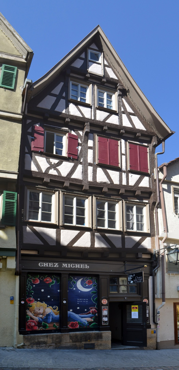 Altes Gebäude mit roten Fensterläden, mehreren Fenstern und einem klassischen Design, das sich auf einer Straße mit einem klaren blauen Himmel im Hintergrund befindet.