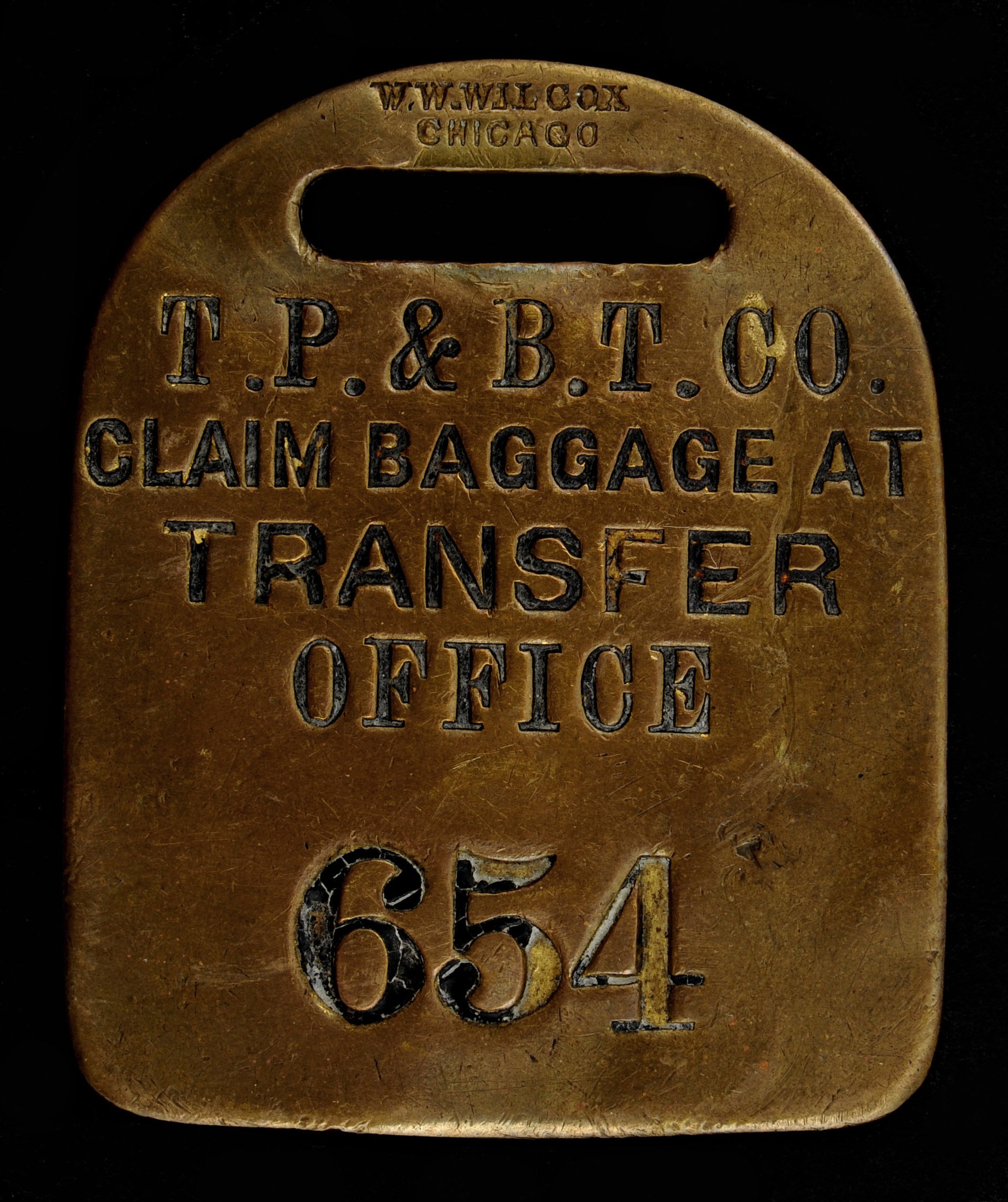 Brass luggage tag mit "T.P. & B.T. Co. Claim Baggage at Transfer Office" eingraviert auf dunklem Hintergrund.