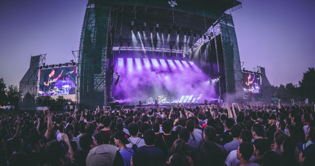 Eine große Menge bei einem Musikfestival schaut auf einer hell erleuchteten Bühne mit Instrumenten und Bildschirmen, die vor einem Gebäude, Bäumen und einem klaren blauen Himmel stehen.