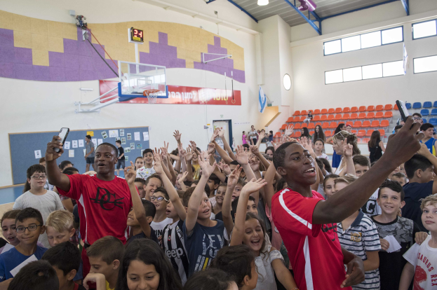 Gruppe von Kindern vor einem Basketballfeld mit Mobiltelefonen, mit einer Anschlagtafel, Uhr, Torpfosten, Basketballkorb, Deckenleuchten, Stühlen und Fenstern im Hintergrund.
