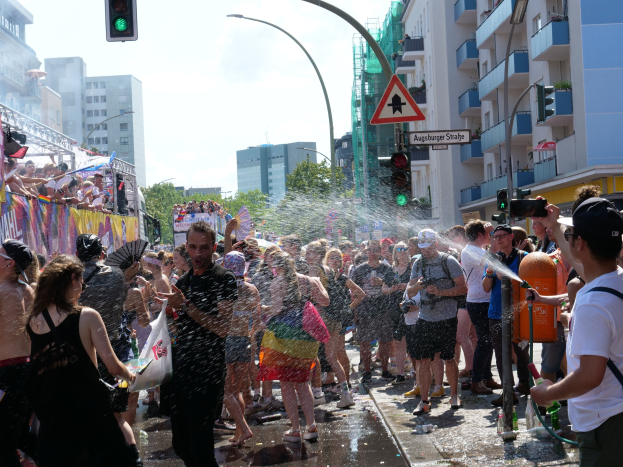 Menschen bei einer Pride-Parade, die sich gegenseitig mit Wasser bespritzen, während sie Gegenstände halten, mit einem Banner links und Gebäuden, Bäumen und Ampeln im Hintergrund.
