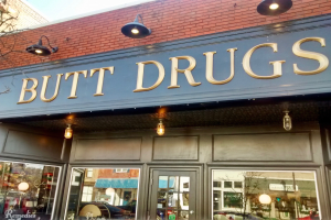 Außenansicht eines Gebäudes mit einer beleuchteten Tafel, auf der "drogen" steht, mit Glaswänden und -türen, umgeben von Fahrzeugen, Gebäuden, Bäumen und Himmel.