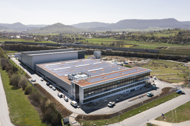 Luftbild einer großen Halle mit Solarmodulen auf dem Dach, umgeben von Fahrzeugen, Bäumen, Gras und Strommasten, mit Hügeln und einem klaren blauen Himmel im Hintergrund; die Halle ist Deutschlands erste solarbetriebene Fabrik.