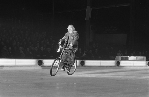 Schwarzes und weißes Foto eines Mannes, der auf einem Eisstadion Fahrrad fährt, mit einer Gruppe von sitzenden Menschen und einer Wand im Hintergrund.
