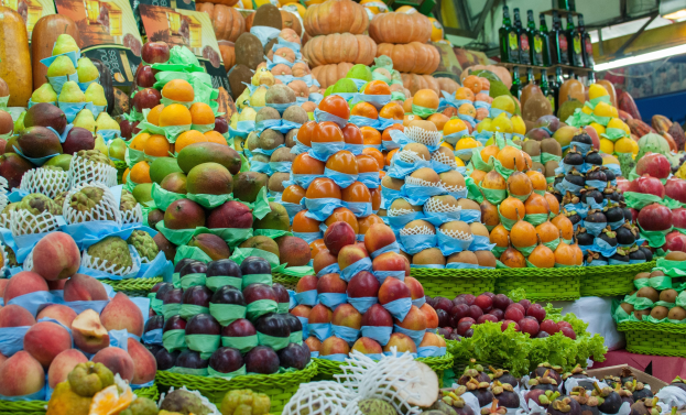 Marktstand mit Äpfeln, Orangen, Birnen und anderen Obst und Gemüse, mit Flaschen auf Regalen und Deckenbeleuchtung.