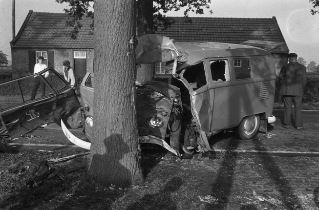 Schwarz-weißes Bild eines demolierten Autos am Straßenrand mit einem Baum im Vordergrund, einigen Menschen in der Nähe und Häusern, Bäumen und einem Mast im Hintergrund.