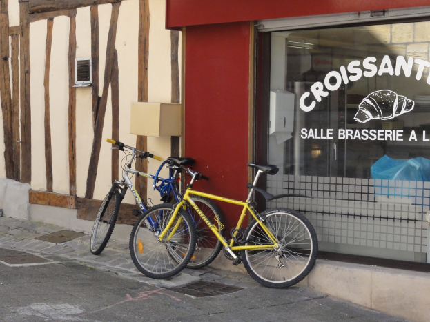 Zwei Fahrräder nebeneinander auf einem Gehweg vor einem Geschäft mit einer Glaswand, auf der Text steht und ein Objekt links von den Fahrrädern zu sehen ist.
