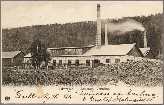 Altes Schwarz-Weiß-Foto einer Fabrik mit Rauch aus Schornsteinen, umgeben von Häusern, Grünflächen und einem Zaun, mit Text unten.